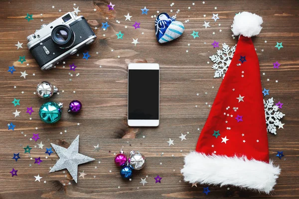 Fondo de Navidad y Año Nuevo con smartphone, cámara antigua, sombrero de Santa Claus rojo, decoraciones: bolas, estrellas, copos de nieve brillantes de plata, confeti en mesa de madera — Foto de Stock