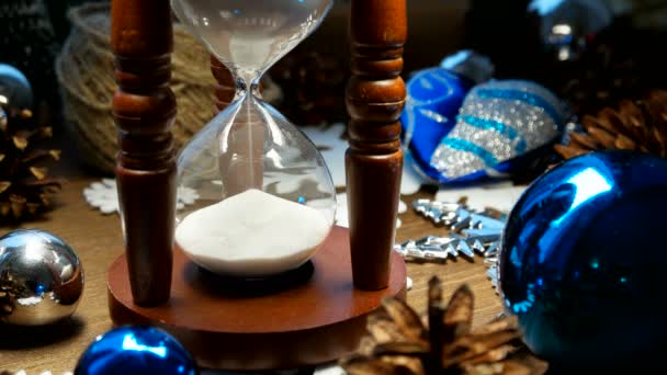 Різдвяний та новорічний фон з подарунками, стрічками, кульками та різними зеленими прикрасами на дерев'яному фоні. Дерев'яний пісочний годинник вимірює час до свята . — стокове відео
