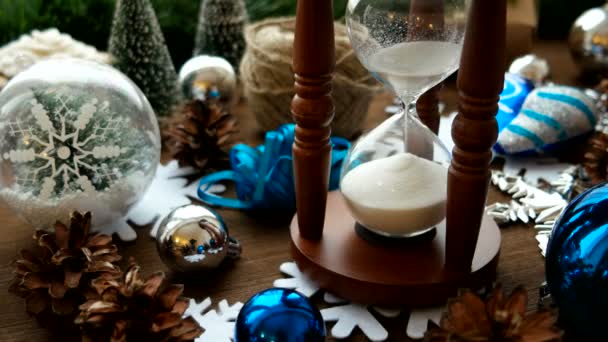 Weihnachten und Neujahr Hintergrund mit Geschenken, Bändern, Kugeln und verschiedenen grünen Dekorationen auf Holzgrund. Holzsanduhr misst Zeit bis zum Urlaub. — Stockvideo