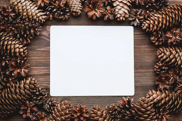 Rustic wooden background with clear sheet of paper and frame of pine cones. Mock up, top view, flat lay. Place for text.