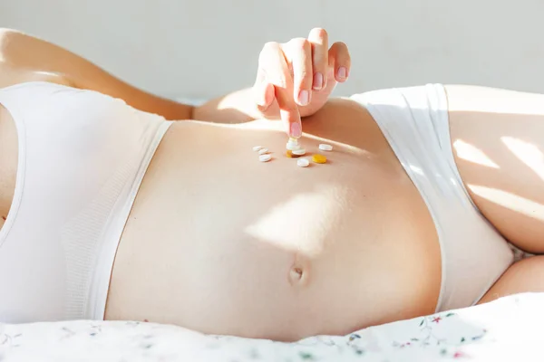 Mujer embarazada en ropa interior blanca acostada en la cama con muchas pastillas. Mujer joven que espera un bebé y tiene que tomar medicamentos . — Foto de Stock