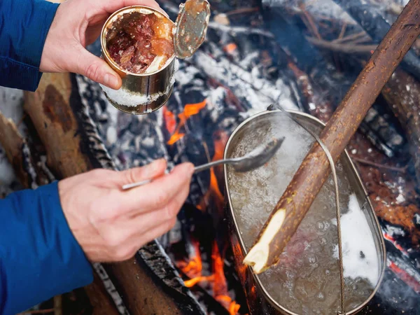Cooking soup on a fire pot. Tourist puts stewed meat in soup from tin can. Winter camping in forest.