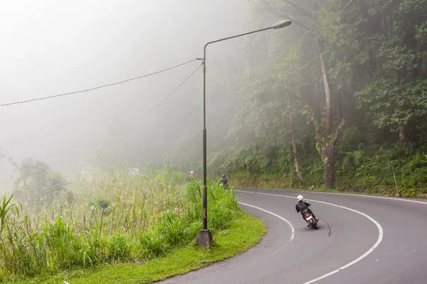 Heavy fog on the road, bad weather for driving motorbike. Road through misty evergreen jungle forest. Winter rainy season. Indonesia.