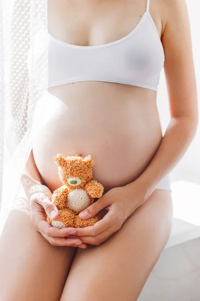 Zwangere vrouw in wit ondergoed met gebreide speeltje teddy beer. Jonge vrouw die een baby verwacht. Gezellige gelukkig achtergrond in zonnige ochtend. — Stockfoto