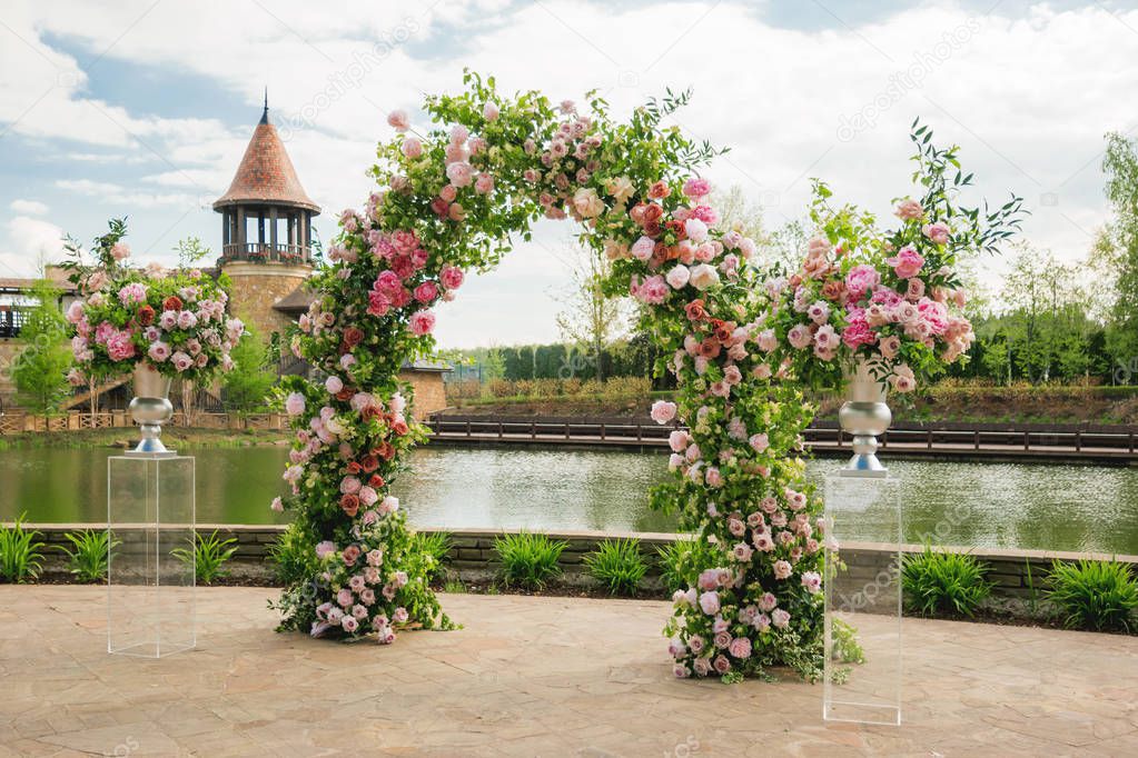 Beautiful floral arch for wedding ceremony. Vases with pink roses and peonies. Wedding set up outdoors in park near pond.