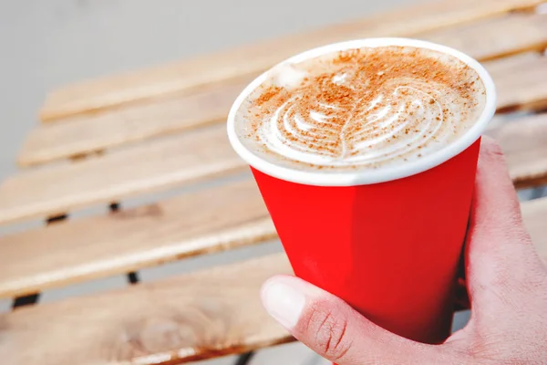 Mujer sosteniendo una taza de papel rojo con café. Café para llevar. Sabrosa bebida caliente en mesa de madera en el día soleado. Comida al aire libre . —  Fotos de Stock