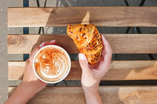 Vrouw met een papieren kopje met koffie en chocolade croissant. Koffie te gaan. Lekkere warme drank op houten tafel in zonnige dag. Buiten de maaltijd. Plat lag, top uitzicht. — Stockfoto