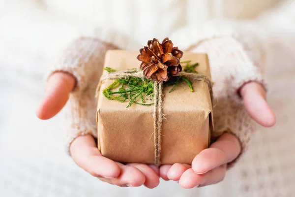 Donna in maglia con un regalo in mano. Il regalo è confezionato in carta artigianale con pigne e legato con corda ruvida. Esempio di modo fai da te per avvolgere un regalo. Posto per il testo . — Foto Stock