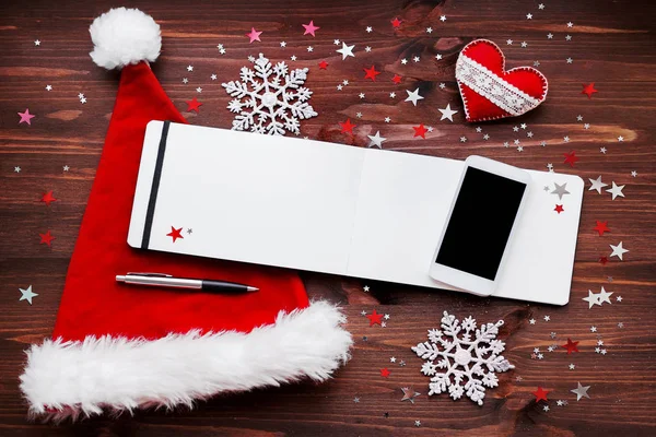 Fondo de Navidad y Año Nuevo con smartphone, sombrero rojo de Santa Claus, bloc de notas con decoraciones de plumas y navidades — Foto de Stock