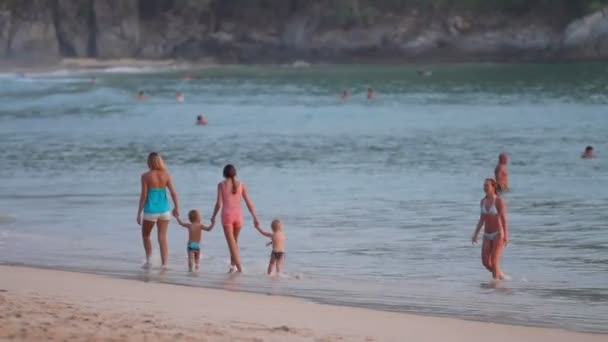 Phuket, Thailand - 18 November 2012. Twee moeders met kinderen lopen op strand. Toeristen zwemmen in zee op zonsondergang. — Stockvideo