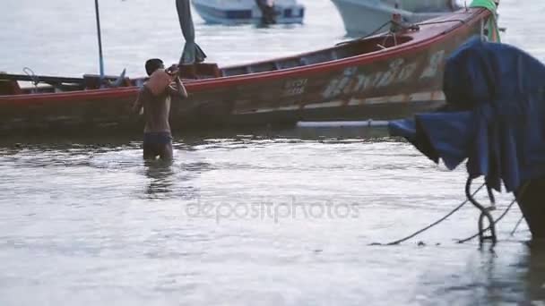Phuket, Thaiföld - November 20, 2012. Napkelte Rawai beach. Seascape halászok csónakokkal. Férfi táska vízben gyaloglás. — Stock videók