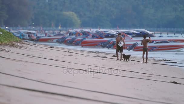 PHUKET, TAILANDIA - 20 de noviembre de 2012. Salida del sol en la playa de Rawai. Gente y perro paseando pasan fila de lanchas amarradas . — Vídeos de Stock