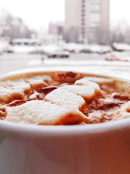Kopje warme cappuccino koffie, stedelijke achtergrond. Smakelijke drank voor raam. Winter straat met gebouwen en auto's op de achtergrond. — Stockfoto