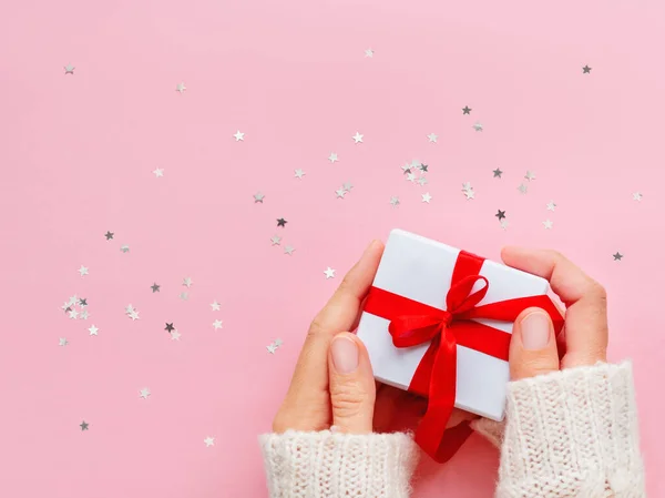 Mujer sostiene un regalo en papel de regalo blanco sobre fondo rosa con confeti estrella brillante. Regalo navideño con cinta roja y lazo. Copiar espacio . —  Fotos de Stock