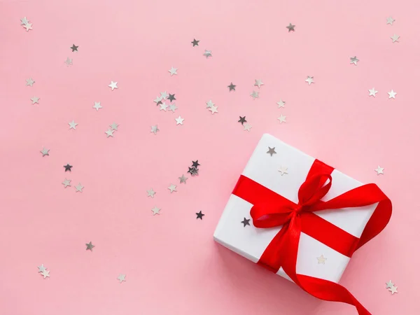 Presente en papel de regalo blanco sobre fondo rosa con confeti estrella plateada. Regalo navideño con cinta roja y lazo. Vista superior, plano . —  Fotos de Stock