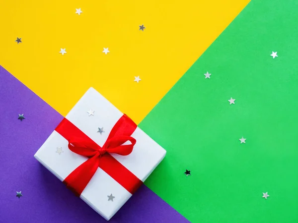 Presente en papel de regalo blanco sobre fondo colorido. Fondo geométrico amarillo, violeta y verde con confeti estrella. Regalo de vacaciones con cinta roja y lazo . —  Fotos de Stock