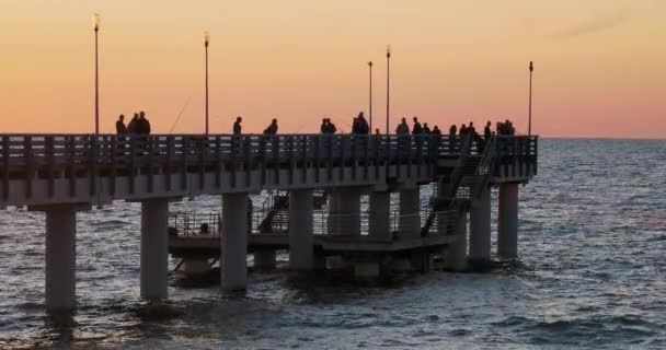 Silhouette di persone che camminano sul molo contro il tramonto. La gente del posto pesca, i turisti camminano prima di andare a letto. Zelenogradsk, Russia . — Video Stock