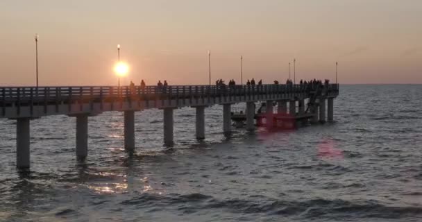 Gün batımına karşı iskelede yürüyen insanların siluetleri. Yerel halk balık tutuyor, turistler yatmadan önce yürüyorlar. Zelenogradsk, Rusya. — Stok video