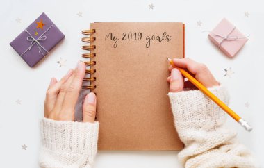 Kadın 2019 golünü kağıt sayfalarla not defterine yazmaya hazırlanıyor. Pembe ve menekşe ambalaj kağıtlarında hediyeler ve arka planda gümüş yıldız konfetileri.