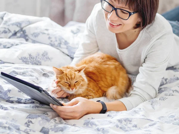 Милые рыжая кошка и женщина в очках лежат в постели. Женщина держит планшет и гладит пушистого питомца. Утренний сон . — стоковое фото