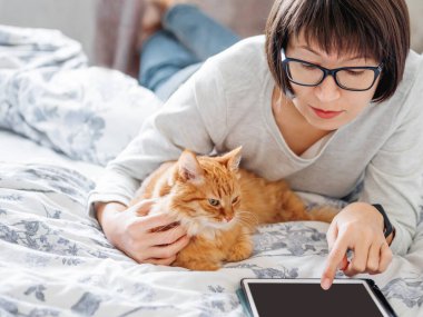 Tatlı zencefilli kedi ve gözlüklü kadın yatakta yatıyor. Kadın tableti tutuyor ve tüylü hayvanını okşuyor. Sabah yatma vakti..