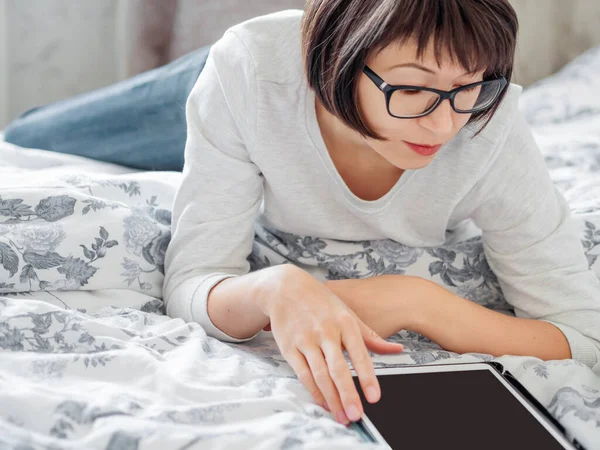 Женщина в очках лежит в постели с планшетом. Она трогает экран. Утренний сон . — стоковое фото