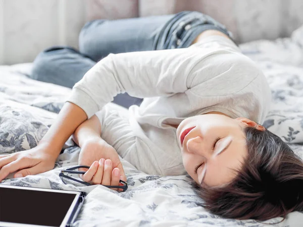 Женщина в очках лежит в постели с планшетом. Она устала и спит после работы онлайн. Утренний сон . — стоковое фото