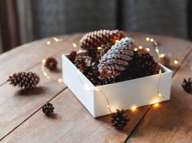 Üzerinde çam kozalakları ve ahşap masa üzerinde ampuller olan beyaz bir kutu. Scandy tarzı. Noel ve Yeni Yıl kutlamaları için hazırlık.
