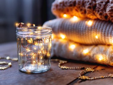 İçinde ampuller olan cam kavanoz. Beyaz, bej ve kahverengi örülmüş süveter yığınının önünde eski püskü ahşap arka planda hoş dekorasyonlar..