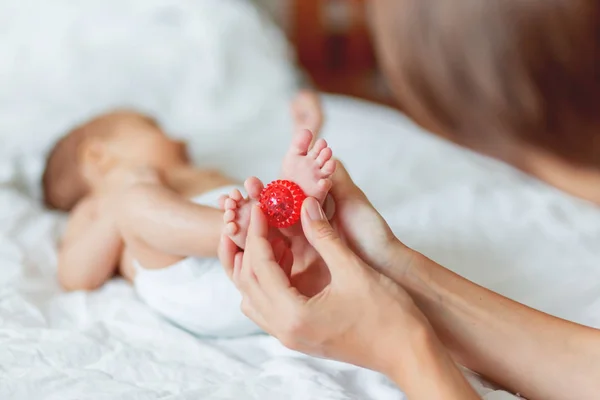 Mother holds newborn baby's feet. Tiny fingers and red massage ball in woman's hand. Cozy morning at home. — Stock Photo, Image