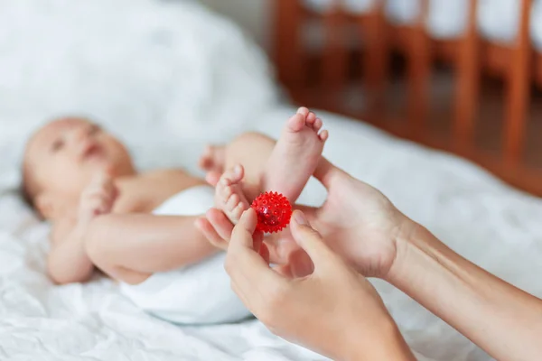Matka trzyma noworodka stopy. Małe palce i Czerwona piłka do masażu w dłoni kobiety. Przytulny poranek w domu. — Zdjęcie stockowe