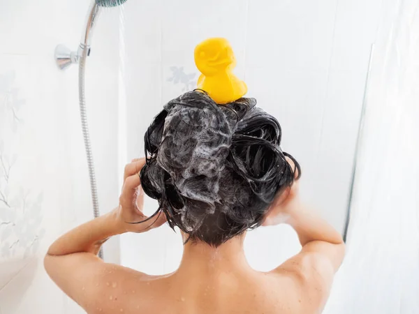 Mulher nua com cabelo curto toma um banho. A mulher lava o cabelo com champô. Pato de borracha amarelo na cabeça da menina no banheiro branco . — Fotografia de Stock