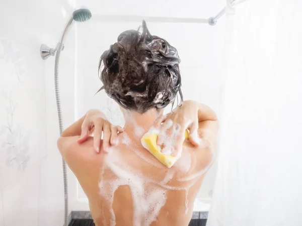 Naked woman with short hair takes a shower. Woman washes her shoulders with yellow sponge. White bathroom. — Stock Photo, Image