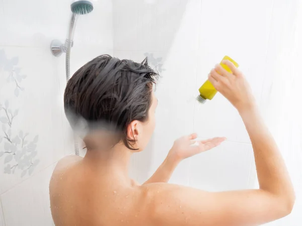 Naga kobieta z krótkimi włosami bierze prysznic. Kobieta wyciska szampon z zielonej butelki. Biała łazienka. — Zdjęcie stockowe