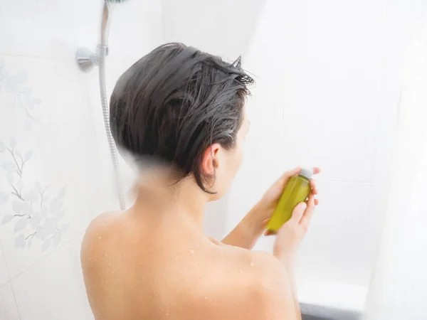 Mulher nua com cabelo curto toma um banho. A mulher está a olhar para a garrafa verde de champô. Banheiro branco . — Fotografia de Stock