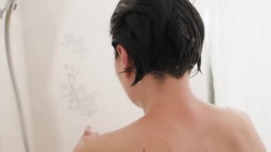 Naked woman takes a shower. Woman washes her short hair with shampoo. Slow motion video in white bathroom.