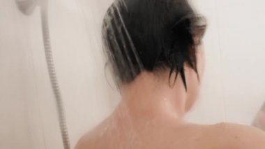 Naked woman takes a shower. Woman washes her short hair with shampoo. Slow motion video in white bathroom.
