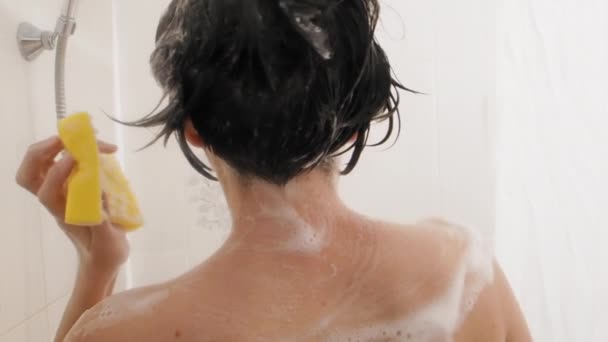 Naked woman with short hair takes a shower. Woman washes her shoulders with yellow sponge. White bathroom. — ストック動画