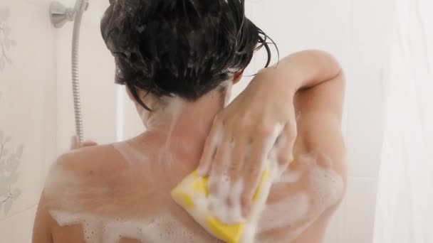 Naked woman with short hair takes a shower. Woman washes her shoulders with yellow sponge. White bathroom. — Stockvideo