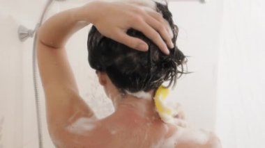 Naked woman with short hair takes a shower. Woman washes her shoulders with yellow sponge. White bathroom.