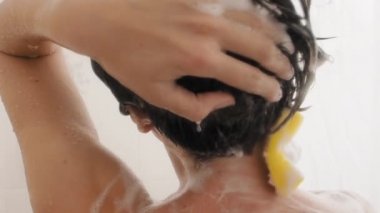 Naked woman with short hair takes a shower. Woman washes her shoulders with yellow sponge. White bathroom.
