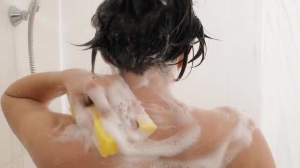 Naked woman with short hair takes a shower. Woman washes her shoulders with yellow sponge. White bathroom. — Stock Video