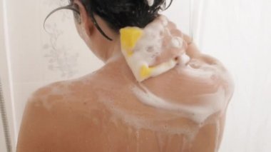 Naked woman with short hair takes a shower. Woman washes her shoulders with yellow sponge. White bathroom.
