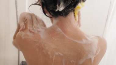 Naked woman with short hair takes a shower. Woman washes her shoulders with yellow sponge. White bathroom.