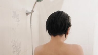Naked woman takes a shower. Woman washes her short hair with shampoo. Slow motion video in white bathroom.