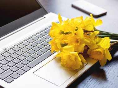 Gümüş metal dizüstü bilgisayarda duran bir buket Narcissus ya da nergis. Taşınabilir cihazda parlak sarı çiçekler. Ahşap arka planda akıllı telefon.