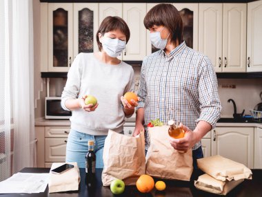 Koruyucu maskeli genç çift mutfaktaki alışverişleri hallediyor. El işi kağıdından yapılmış poşetlerdeki ürünler. Coronavirus COVID19 nedeniyle karantina koşullarında yiyecek teslimatı.
