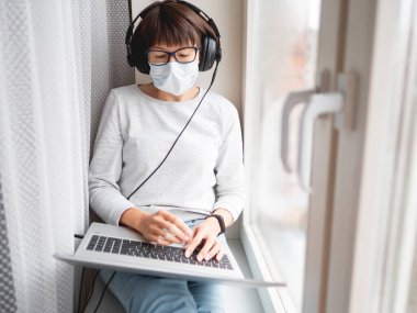 Tıbbi maskeli kadın uzaktan kumanda ile evden çalışıyor. Diz üstü bilgisayarı ve kulaklığı ile pencere pervazında oturuyor. Coronavirüs COVID19 yüzünden karantina altına alındı. Evde yalnızlık.