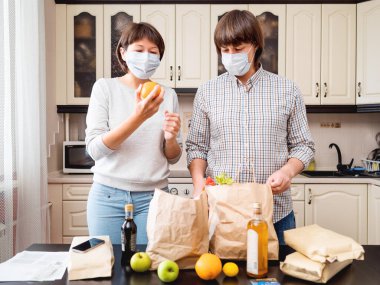 Koruyucu maskeli genç çift mutfaktaki alışverişleri hallediyor. El işi kağıdından yapılmış poşetlerdeki ürünler. Coronavirus COVID19 nedeniyle karantina koşullarında yiyecek teslimatı.