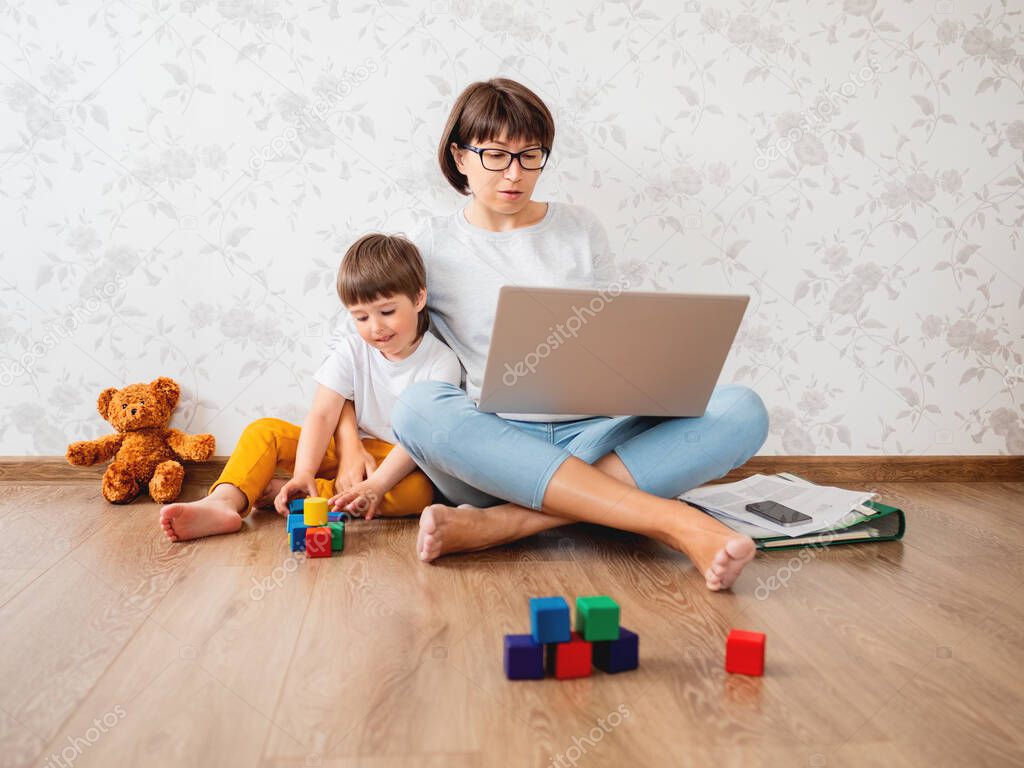 Mom and son sitting at home quarantine because of coronavirus COVID19. Mother remote works with laptop, son plays with toy blocks. Self isolation at home.